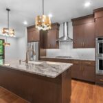7736 Turnberry Kitchen. Granite Countertops. Modern Slab Front Stained Maple Cabinets. Gold Hardware. Wall Oven. Tiled Backsplash. Chimney Range Hood.