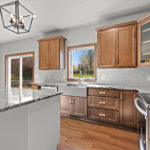 3320 Cottage Hill Kitchen. Stained Maple Perimeter Cabinets with Painted White Island. Farmhouse Apron-Front Sink. Granite Countertops. Tiled Backsplash. Luxury Vinyl Plank Floors.