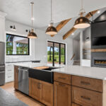 704 Royal St Pats Kitchen. Painted Perimeter Cabinets with Stained Maple Island and Custom Wood Hood. Pendant Lights. Under-Counter Beverage Center Refrigerator. Interior Transom. White Vertical Picket Tiled Backsplash. Glass Doors Above Upper Cabinets. Calacatta Prado Quartz with Black Pearl Brushed Granite Countertops. Black Painted Shiplap (Sherwin-Williams Iron Ore SW-7069) Fireplace Surround with Mitered Corners and Reclaimed Hewn Timber Mantel. Stained Maple Floating Shelves. TV Above Fireplace. Luxury Vinyl Plank Flooring. Hewn Timber Ceiling Beams at Vaulted Ceiling. Black Windows with White Painted Trim.