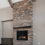 2966 Sugar Bush Great Room + Fireplace. Eldorado "Mountain Ledge Bitter Root" stone fireplace surround with steel mantel. Knotty alder stained cabinets. Cathedral ceiling with window wall and hewn timber ceiling beams. Adura Rigid Plank "Dockside Drift" luxury vinyl plank floors.