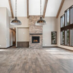 2966 Sugar Bush Great Room + Fireplace. Eldorado "Mountain Ledge Bitter Root" stone fireplace surround with steel mantel. Knotty alder stained cabinets. Cathedral ceiling with window wall and hewn timber ceiling beams. Adura Rigid Plank "Dockside Drift" luxury vinyl plank floors.