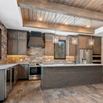 2966 Sugar Bush Kitchen. Eldorado "Mountain Ledge Bitter Root" backsplash wall stone. Knotty alder stained cabinets with wide plank island. Tray ceiling with weathered shiplap planks and hewn timber beams. Stoll black riveted steel custom range hood. Adura Rigid Plank "Dockside Drift" luxury vinyl plank floors. Lennon leathered granite countertops. Under-counter beverage centers. Reclaimed barn wood sliding barn door.