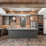2966 Sugar Bush Kitchen. Eldorado "Mountain Ledge Bitter Root" backsplash wall stone. Knotty alder stained cabinets with wide plank island. Tray ceiling with weathered shiplap planks and hewn timber beams. Stoll black riveted steel custom range hood. Adura Rigid Plank "Dockside Drift" luxury vinyl plank floors. Lennon leathered granite countertops. Under-counter beverage centers. Reclaimed barn wood sliding barn door.