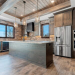 2966 Sugar Bush Kitchen. Eldorado "Mountain Ledge Bitter Root" backsplash wall stone. Knotty alder stained cabinets with wide plank island. Tray ceiling with weathered shiplap planks and hewn timber beams. Stoll black riveted steel custom range hood. Adura Rigid Plank "Dockside Drift" luxury vinyl plank floors. Lennon leathered granite countertops.