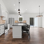 7730 Prestwick Kitchen & Dining. White Painted Woodwork & Cabinetry. Black Granite Countertops. Luxury Vinyl Plank Floors. Slide-In Electric Range. Tiled Backsplash. Island with Paneled Ends & Pendant Lights Above. White Painted Wood Range Hood Cover.