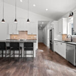 7730 Prestwick Kitchen. White Painted Woodwork & Cabinetry. Black Granite Countertops. Luxury Vinyl Plank Floors. Slide-In Electric Range. Tiled Backsplash. Island with Paneled Ends & Pendant Lights Above. White Painted Wood Range Hood Cover.