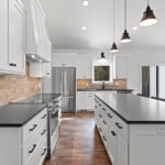 7730 Prestwick Kitchen. White Painted Woodwork & Cabinetry. Black Granite Countertops. Luxury Vinyl Plank Floors. Slide-In Electric Range. Tiled Backsplash. Island with Paneled Ends & Pendant Lights Above. White Painted Wood Range Hood Cover.